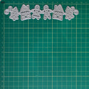 A set of five silver metal cookie cutters shaped like gingerbread houses, trees, and a gingerbread person, arranged in a row on a green cutting mat with a grid.