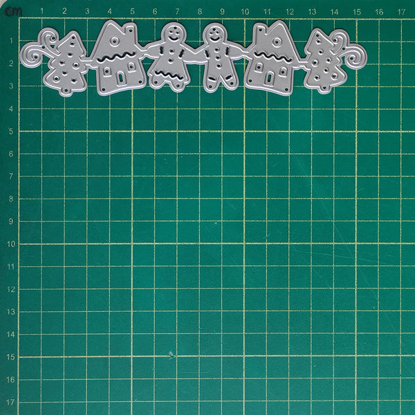 A set of five silver metal cookie cutters shaped like gingerbread houses, trees, and a gingerbread person, arranged in a row on a green cutting mat with a grid.