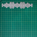 A silver metal die-cut ornament or decorative trim piece with intricate snowflake and scroll patterns, laid horizontally on a green cutting mat with grid lines.
