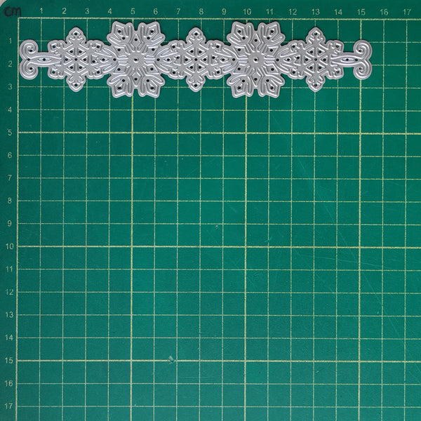 A silver metal die-cut ornament or decorative trim piece with intricate snowflake and scroll patterns, laid horizontally on a green cutting mat with grid lines.