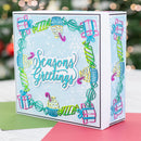 A square greeting card or gift box with a festive candy-themed border and the words 'Seasons Greetings' on the front, photographed on a colorful surface with a blurred holiday background.