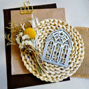A silver metal gothic arch window cutout ornament displayed on a round woven straw mat, with dried flowers and a kraft paper clipboard in the background.