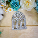 A metal die-cut craft embellishment shaped like a Gothic-style arched window, displayed on a beige textured fabric background with decorative flowers.