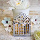 A silver metal cookie cutter with an arched, ornate window design, shown on a rustic wooden surface with decorative white and pastel flowers.