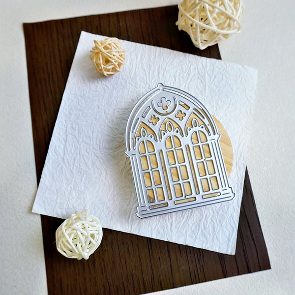 A silver metal die-cut window frame ornament lies on a textured white paper square, placed on a dark brown wood board with decorative cream yarn balls around it.