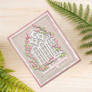 A handmade square greeting card with a pink border, featuring a white arched window design surrounded by pink flowers and green leaves, with the words 'seasons love' in white script. The card is displayed on a light wood surface with green fern leaves framing the scene.