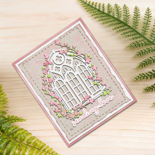 A handmade square greeting card with a pink border, featuring a white arched window design surrounded by pink flowers and green leaves, with the words 'seasons love' in white script. The card is displayed on a light wood surface with green fern leaves framing the scene.