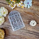 A silver metal arch-shaped decorative frame or tray with intricate scrollwork, displayed on a rustic wooden surface surrounded by artificial flowers and woven decor.