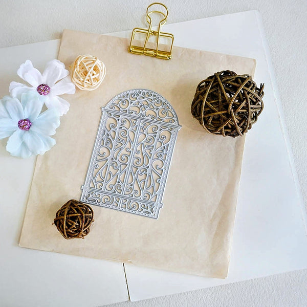 A silver metal die-cut stencil with an ornate scrollwork pattern and arched top, displayed on a beige sheet of craft paper with decorative props including white flowers, woven balls, and a gold binder clip.