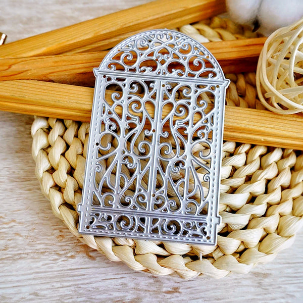 A silver metal die-cut ornament with an ornate filigree gate design, displayed on a woven straw mat with wooden sticks in the background.