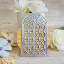 A silver metal die-cut craft embellishment shaped like an arched window with geometric lattice patterns, displayed on a wooden stand with decorative white flowers in the background.