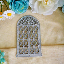 A gray metal die-cut craft stencil shaped like an arched window with ornate scrollwork and geometric lattice patterns, displayed on a beige textured fabric background with decorative flowers.