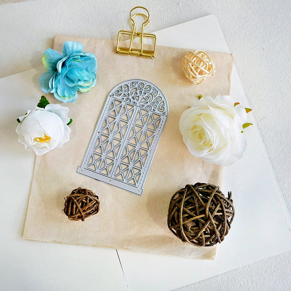 A silver metal die-cut arch window frame template displayed on a beige parchment paper background, surrounded by decorative props including artificial flowers, a gold binder clip, and woven balls on a white surface.