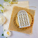 A white arched window-shaped decorative plaque with an intricate lattice design, displayed on a round woven mat over a burlap fabric square, with white flowers and a gold clip in the background.