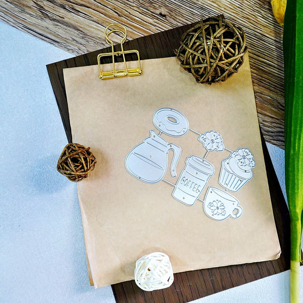 A kraft paper bag with a white line-art coffee-themed illustration, clipped at the top with a gold metal binder clip and arranged on a dark wood surface with decorative woven balls and a green stem.