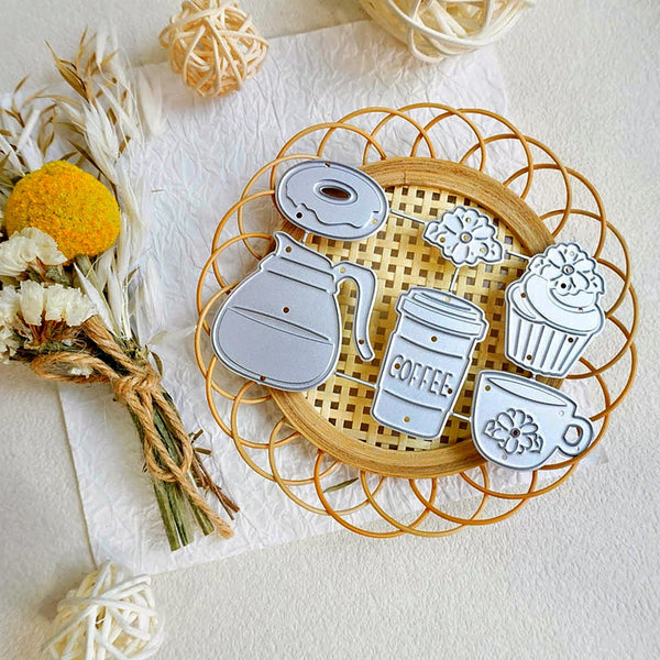 A set of silver metal cookie cutters shaped like coffee and bakery items, including a coffee pot, takeaway coffee cup, donut, cupcake, and coffee cup, displayed on a round woven tray with dried flowers as props.