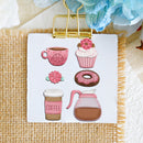 A white rectangular card clipped at the top with a gold binder clip, featuring six coffee-themed illustrations: a pink floral mug, a pink cupcake with flowers, a pink flower patch, a pink-glazed donut, a coffee cup with a pink sleeve, and a pink-and-brown coffee carafe. The card is placed on a beige woven mat with decorative fabric and flowers in the background.