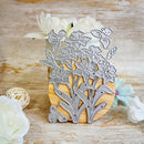 A metal floral die-cut cutting template with daffodils, tulips, and butterflies, displayed on a wooden block with decorative white and blue flowers on a rustic wooden surface.