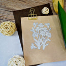 A kraft paper bag clipped at the top with a gold binder clip, featuring a white line-art floral bouquet design printed on it. The bag is styled on a dark wood surface with decorative rattan balls, a green leaf, and a yellow fruit slice.