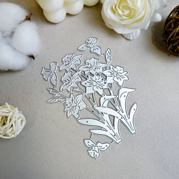 A silver metal floral die-cut cutting template with intricate flower and leaf shapes, displayed on a textured light gray surface with decorative white flowers and cotton props.