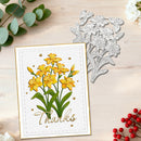 A handmade greeting card with yellow lilies and the word 'Thanks' in gold script, shown on a light wood surface alongside a silver floral die-cutting template.