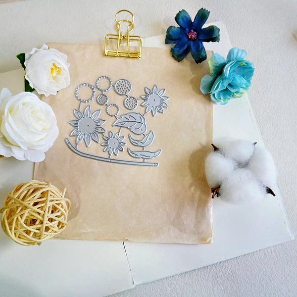 A flat lay of a metal floral die-cutting or scrapbooking template sheet placed on a beige sheet of craft paper, surrounded by decorative props including white and blue flowers, cotton bolls, a yellow woven ball, and a gold binder clip on a white surface.