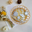 A flat-lay of metal jewelry making dies arranged on a round bamboo tray, with dried flowers and decorative yarn balls as props on a textured cream background.