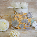 A set of metal floral and leaf-shaped craft dies displayed on a wooden board, with a white pot of white flowers behind it and decorative white flowers on a light wood surface.