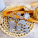 A set of silver metal craft dies for cutting out shapes like suns, leaves, crescent moons, and floral patterns, displayed on a woven straw mat with wooden sticks and decorative balls.