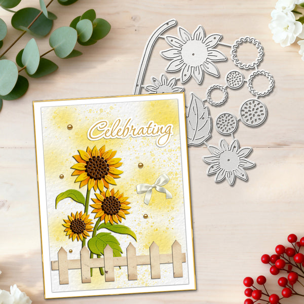 A handmade greeting card with sunflowers, green leaves, and a picket fence, displayed next to metal flower and leaf die-cut templates on a light wood surface with decorative eucalyptus and red berries.