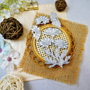 A set of silver metal floral dies arranged on a round wooden hoop over a burlap mat, with decorative flowers and woven balls around it on a light gray surface.