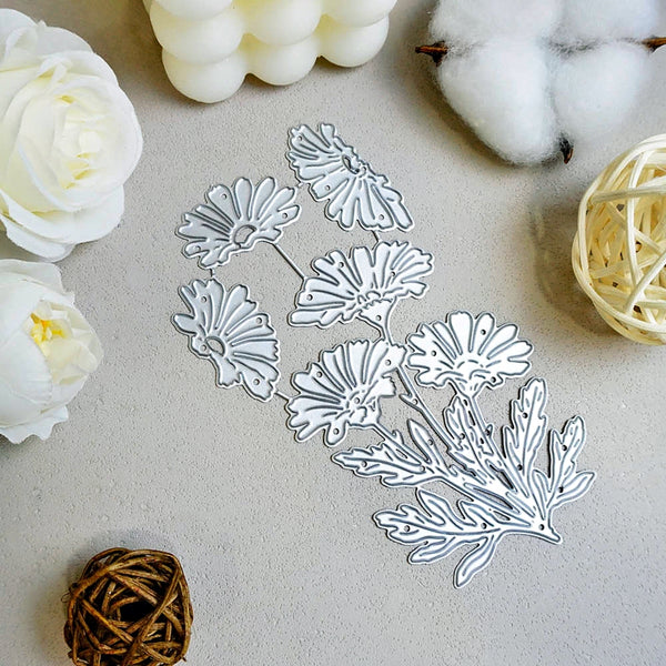 A set of silver metal floral die-cut craft cutters arranged on a light beige textured surface, surrounded by decorative props including white flowers, cotton bolls, and woven balls.