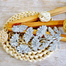 A set of silver metal floral die-cut stickers or decals arranged on a woven straw mat with wooden sticks and small white yarn balls as styling props.