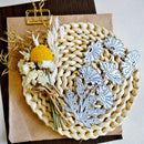 A flat-lay of silver metal floral dies arranged on a round woven straw mat, with dried white flowers, a yellow pom-pom, and a gold binder clip on brown paper against a dark wood surface.