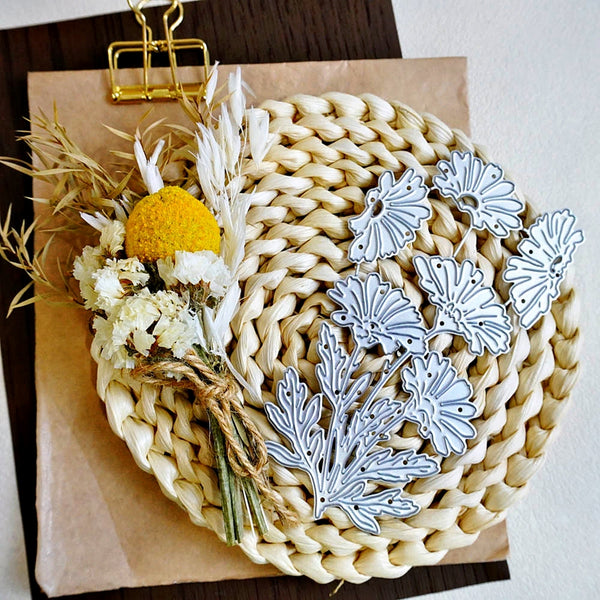A flat-lay of silver metal floral dies arranged on a round woven straw mat, with dried white flowers, a yellow pom-pom, and a gold binder clip on brown paper against a dark wood surface.