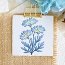 A square white card or small sign featuring an embroidered floral design with three light blue flowers and yellow centers, held at the top by a gold binder clip. The card is placed on a woven beige fabric surface with decorative paper flowers around it.