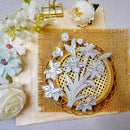 A floral metal die-cut craft stamp set featuring white lilies and daffodils, displayed on a round wooden tray over a burlap mat with decorative white flowers and a gold binder clip.