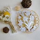A white floral die-cut sticker or decal sheet featuring daffodils and butterflies, displayed on a gold-colored circular wire stand alongside decorative dried flowers, small woven balls, and a textured cream background.