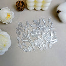A floral metal cutting die sheet with intricate silver-toned leaf and blossom outlines, displayed on a light beige textured surface with decorative white flowers, cotton bolls, and a woven brown sphere around it.