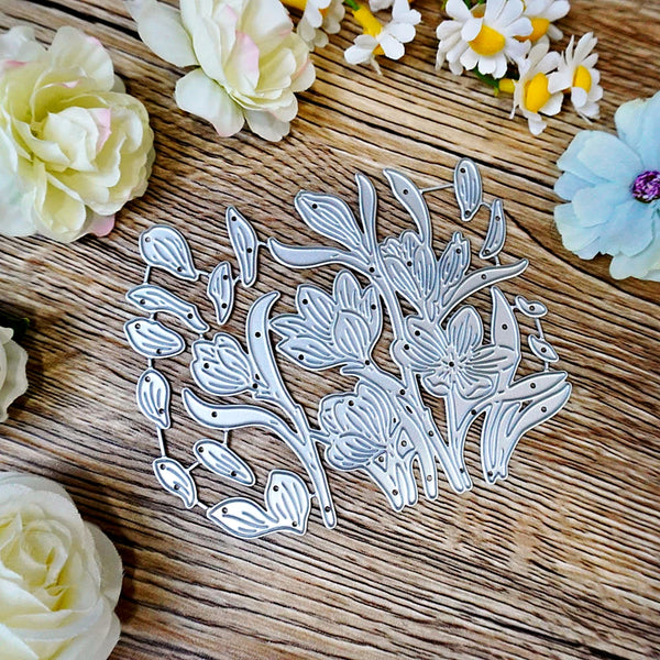 A floral metal die-cut template or craft stencil with layered leaf and flower shapes, displayed on a rustic wooden surface surrounded by decorative artificial flowers.