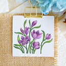A square white card or small sign clipped at the top with a gold binder clip, featuring embroidered purple flowers with green stems on a beige woven fabric background.