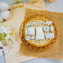 A set of white metal stamp dies arranged on a round woven tray over a burlap mat, with cotton bolls and white flowers as styled props on a light surface.