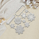 A set of silver metal snowflake and decorative frame-shaped dies arranged on a textured cream fabric with cotton stems and a small wooden tag nearby.