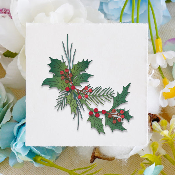 A square white card featuring a layered paper cutout of green holly leaves and pine branches with red berries, styled on a beige fabric background surrounded by white and blue flowers.