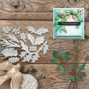 A flat-lay of Christmas-themed crafting supplies on a rustic wooden background, featuring a set of silver metal die-cutting templates for holly and berries alongside a handmade layered card decorated with green holly leaves, red berries, and a black 'Merry Christmas' banner.