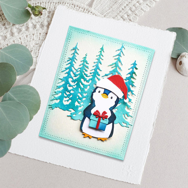 A square handmade greeting card featuring a cute penguin wearing a red Santa hat and holding a blue gift box, set against a snowy pine tree background. The card is displayed on a textured white cardstock sheet with decorative eucalyptus leaves around it.