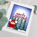 A square Christmas greeting card featuring a cartoon mouse in a red hat and scarf, surrounded by snowy pine trees and a large moon, with the words “merry CHRISTMAS” in red. The card is photographed on a light fabric surface with decorative green leaves around it.