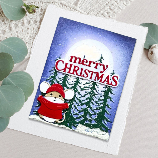 A square Christmas greeting card featuring a cartoon mouse in a red hat and scarf, surrounded by snowy pine trees and a large moon, with the words “merry CHRISTMAS” in red. The card is photographed on a light fabric surface with decorative green leaves around it.