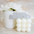 A white textured candle with a bubbled, puffy design sits next to a white ceramic cup filled with white flowers, featuring a silver metal 'Merry Christmas' sign in the foreground, all set on a light wood background.