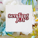 A square white card with torn paper edges featuring the phrase 'sending JOY' in red and white script, surrounded by artificial white, blue, and yellow flowers on a beige fabric background.